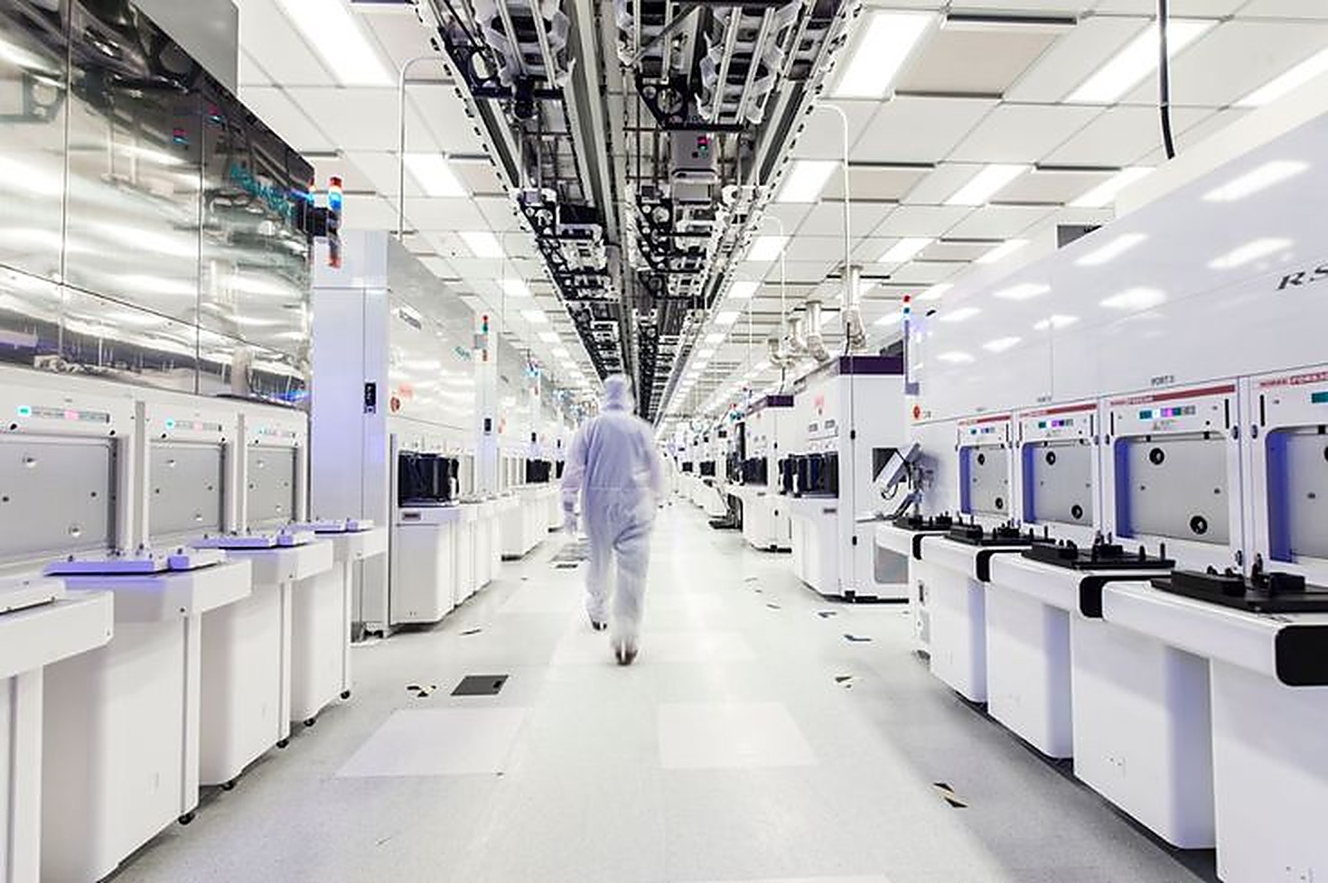 A semiconductor cleanroom: a technician in protective coveralls walking between rows of wafer-processing tools.